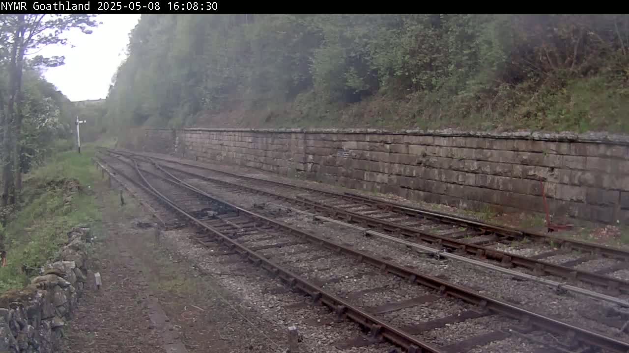 A view of a railway track on an overcast day, with a stone wall to the right and lush green vegetation on both sides.