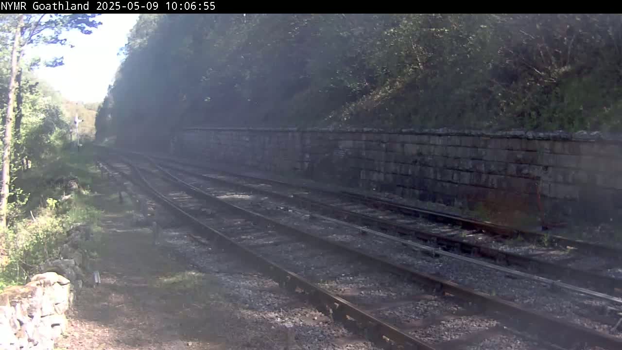 A hazy view of a railway line runs alongside a stone wall, bordered by trees and vegetation under a partly sunny sky.