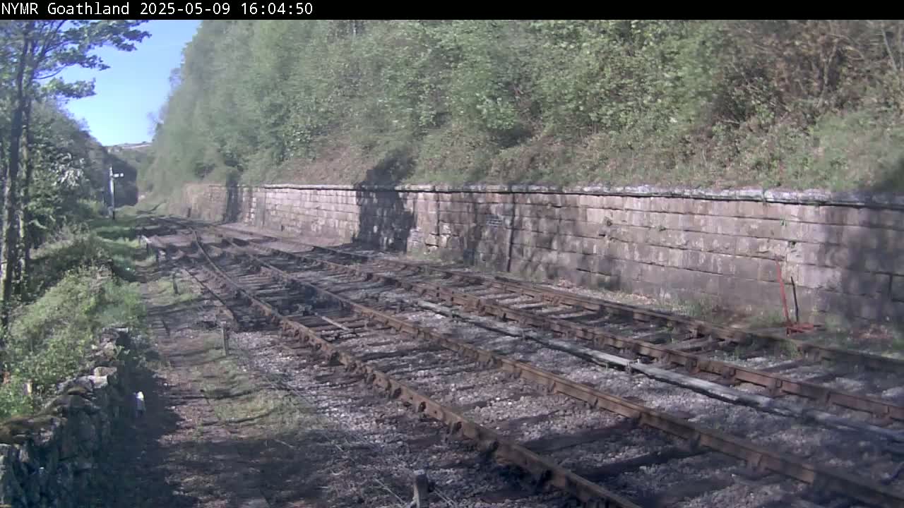A sunny view of railway tracks alongside a stone wall and lush greenery.