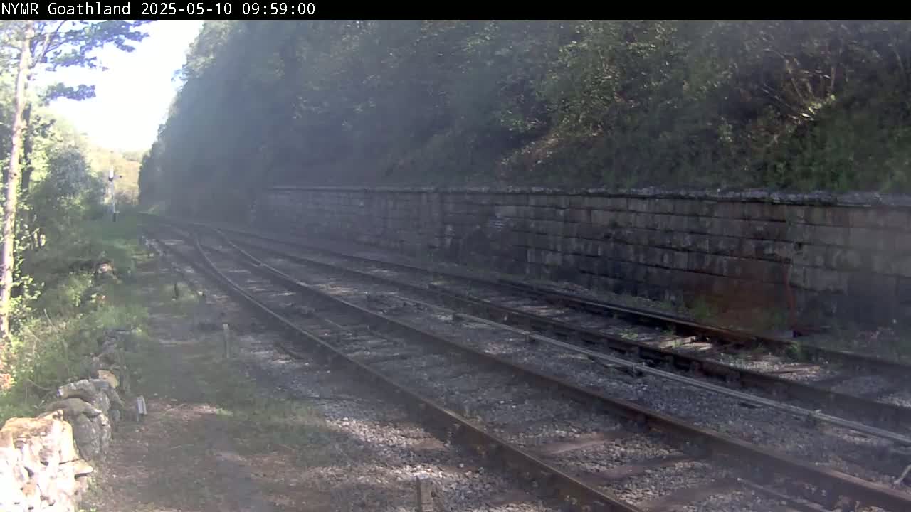 A hazy view of a railway track runs alongside a low stone wall, bordered by trees and greenery under a somewhat bright sky.