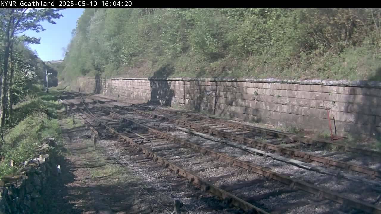 Multiple train tracks curve gently around a stone wall, bordered by lush green vegetation under a sunny sky.