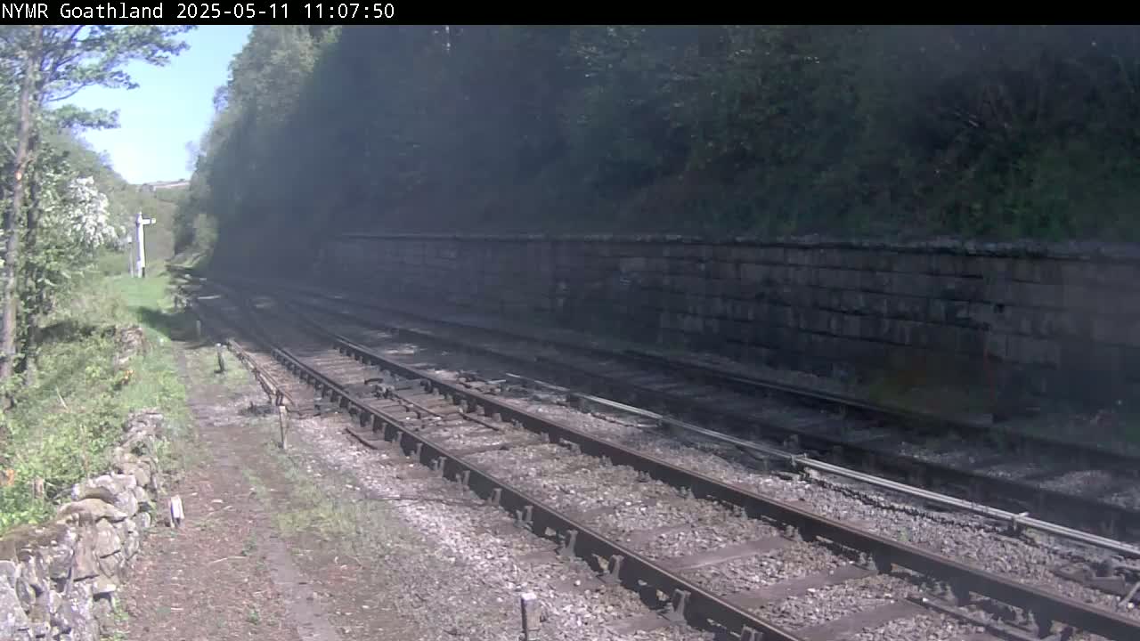 A view of a railway track on a sunny day, with a stone retaining wall alongside, and green vegetation on either side.