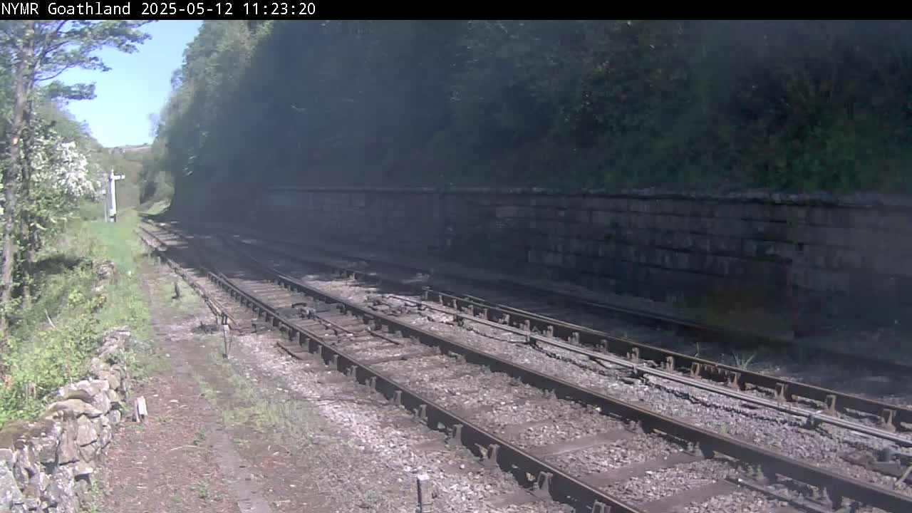 Two sets of train tracks curve gently to the right, passing alongside a stone wall and lush green vegetation under a sunny sky.