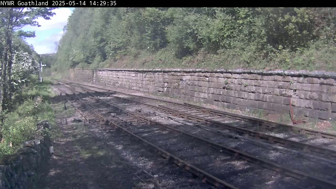 Train tracks run alongside a stone wall, bordered by lush green vegetation under a sunny sky.