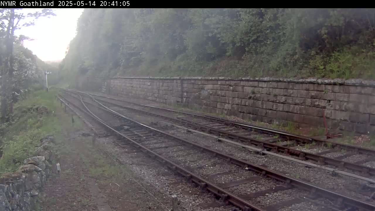 Two sets of train tracks curve through a misty, green landscape alongside a low stone wall.