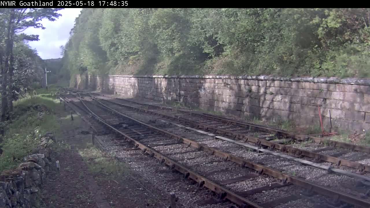 Multiple train tracks curve gently to the left, beside a stone wall and lush green vegetation under a partly cloudy sky.