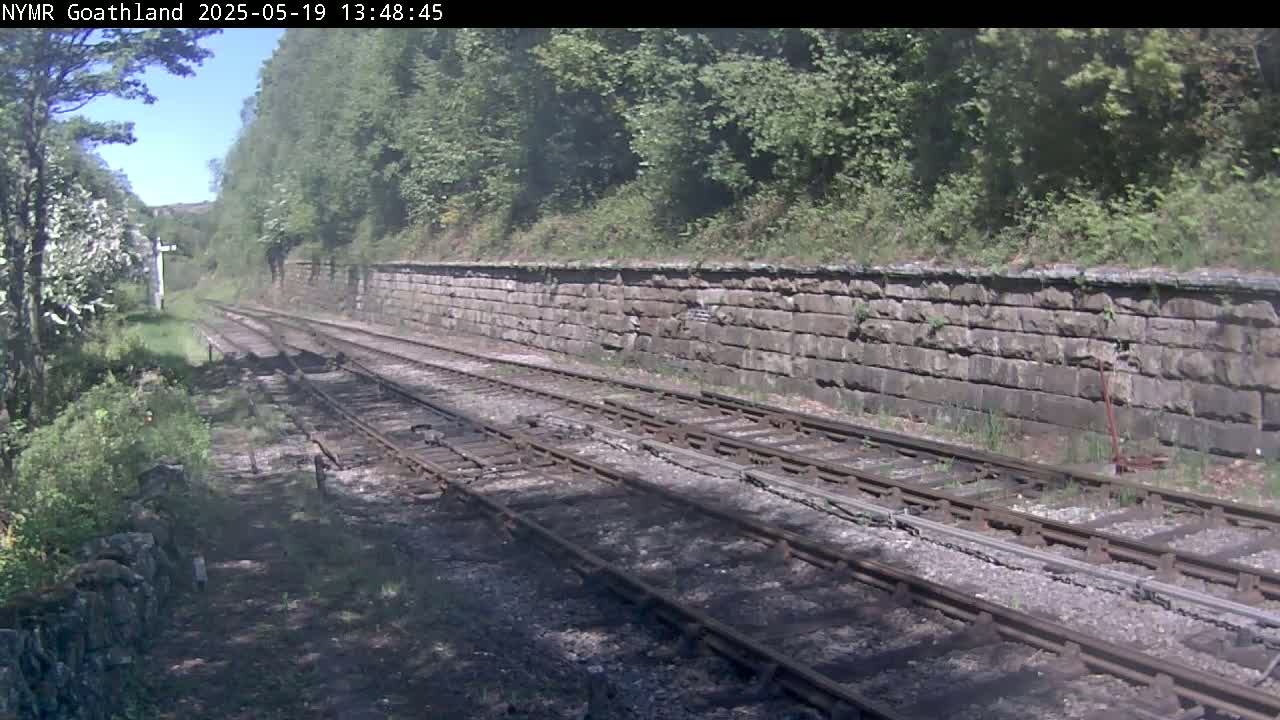 A sunny day reveals a view of railway tracks running alongside a stone wall, bordered by lush green vegetation.