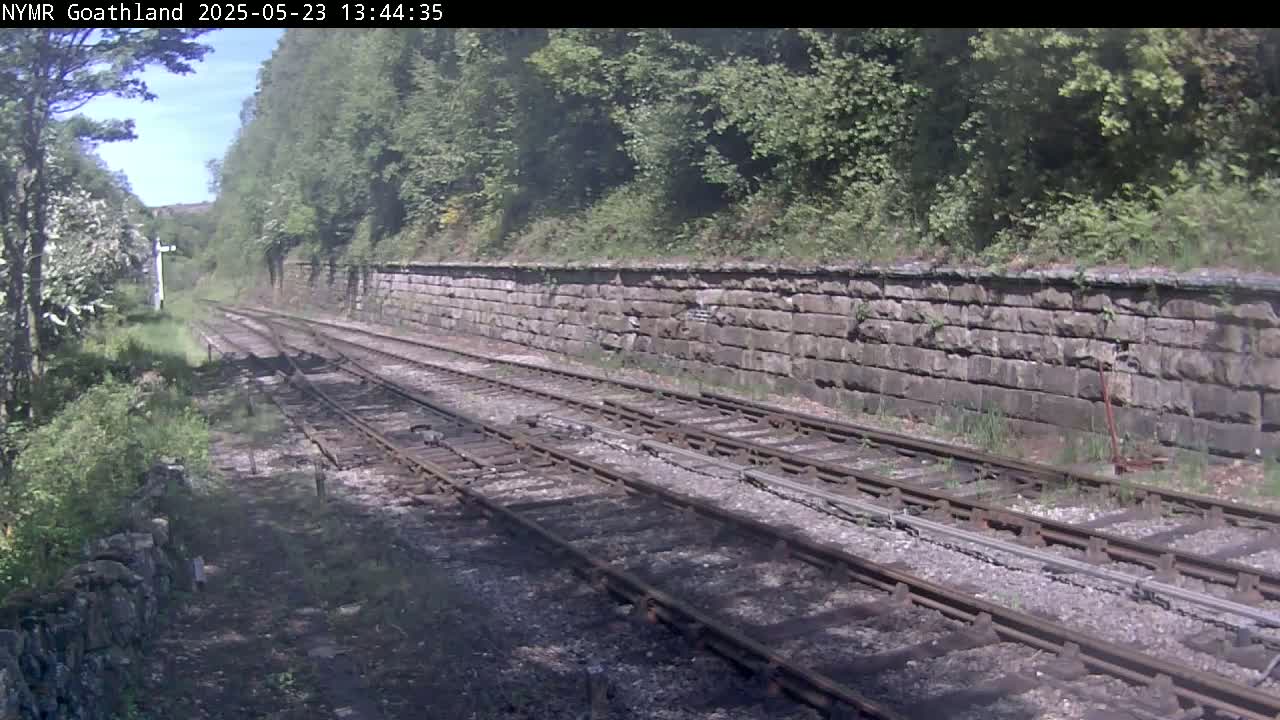 A view down a railway line on a sunny day, bordered by a stone wall and lush green vegetation.