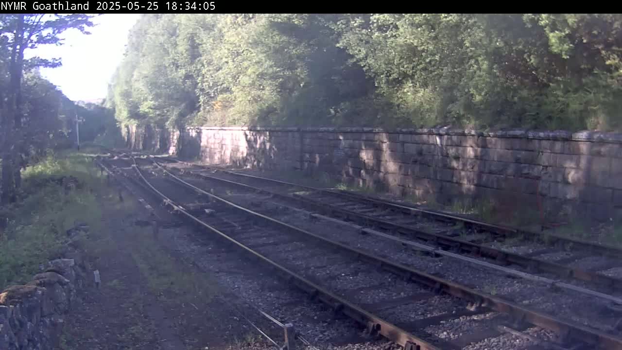 Three sets of train tracks wind through a landscape of lush greenery and stone walls, under a hazy, sunny sky.