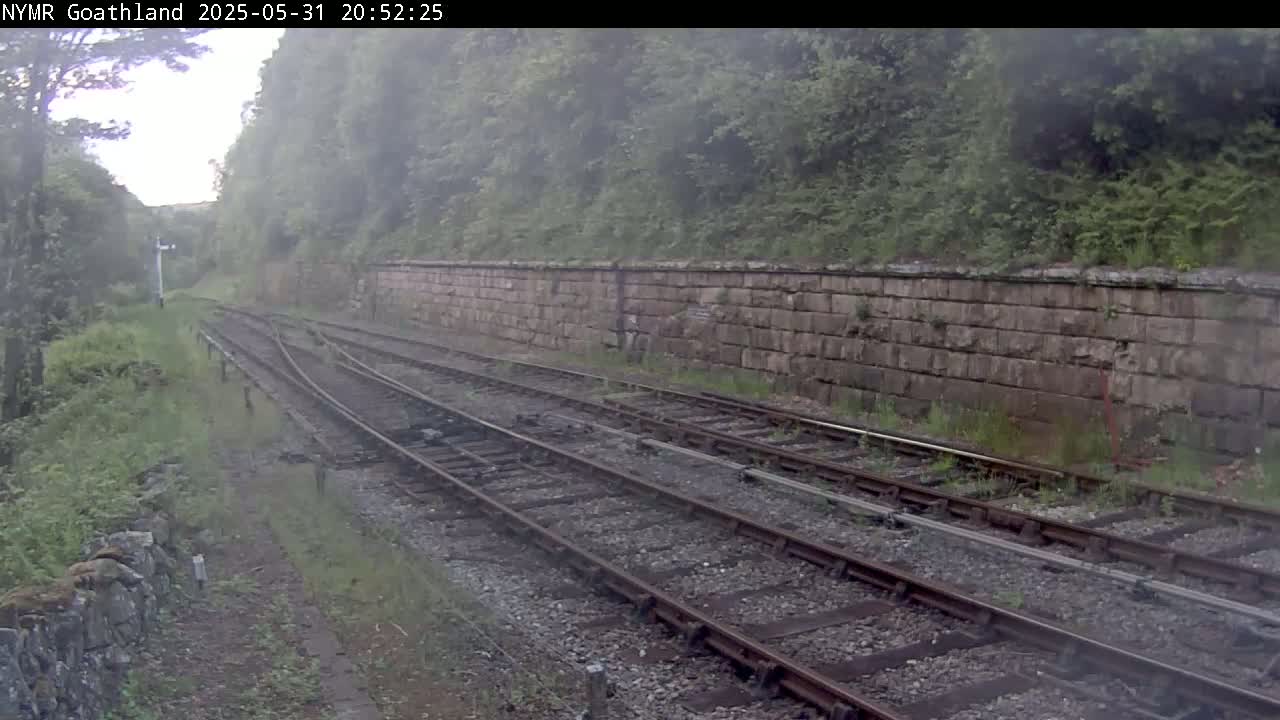A misty view of railway tracks curving alongside a stone retaining wall, bordered by lush green vegetation.