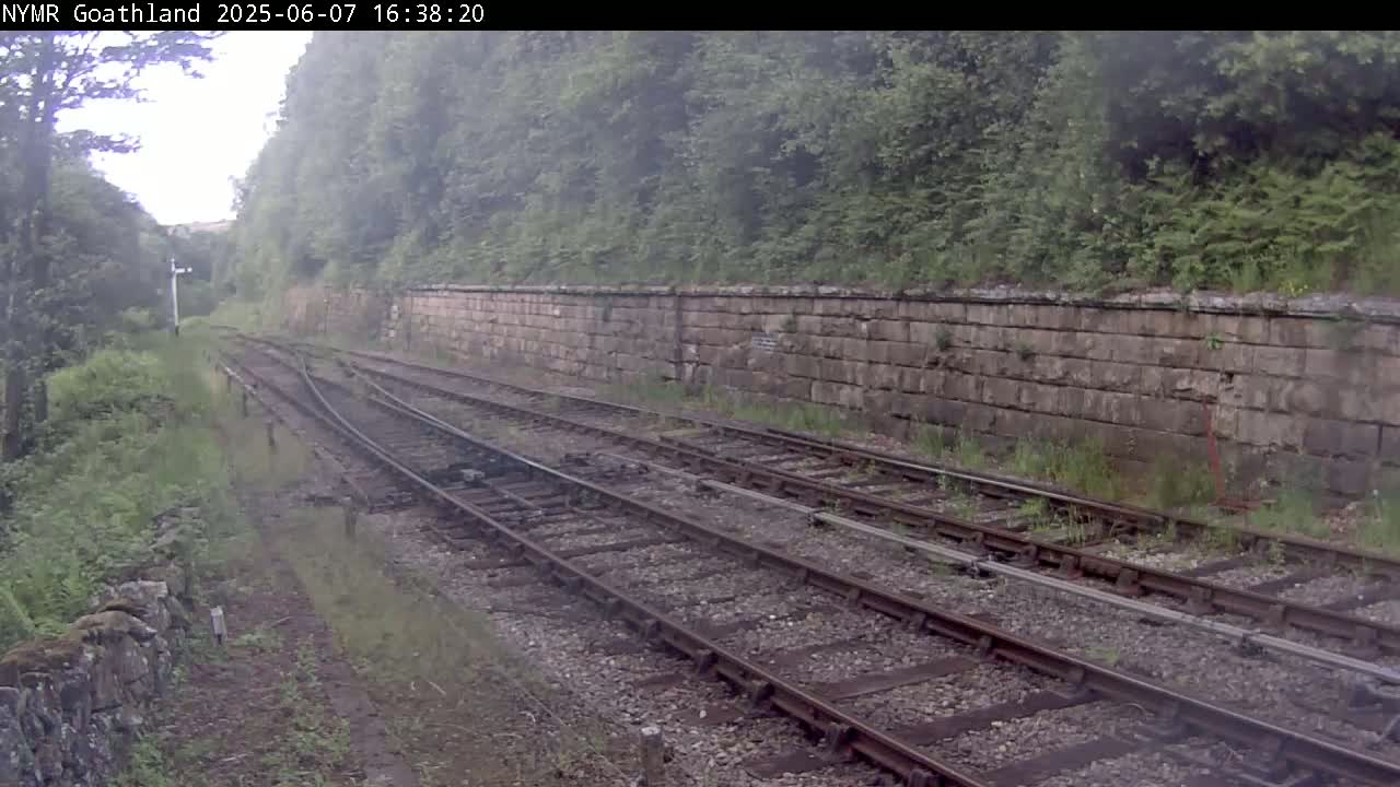 A view of a railway track with a stone wall to the right, surrounded by lush green vegetation on an overcast day.