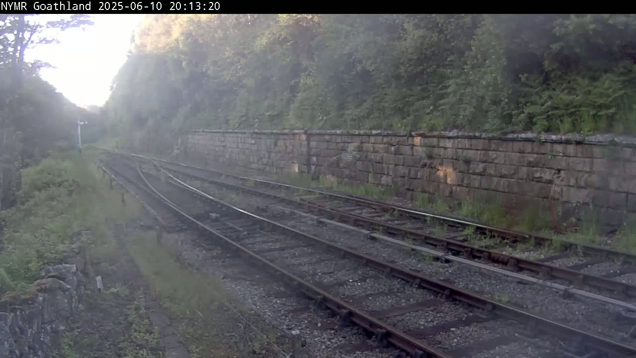 A view of a railway line with two sets of tracks running alongside a stone wall, partially obscured by mist or fog under a hazy sky.