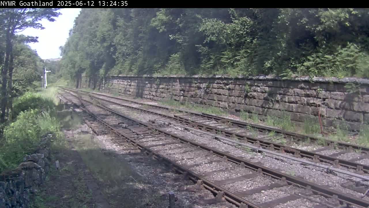 A view of a railway track running between a stone wall and lush green vegetation under a partly cloudy sky.