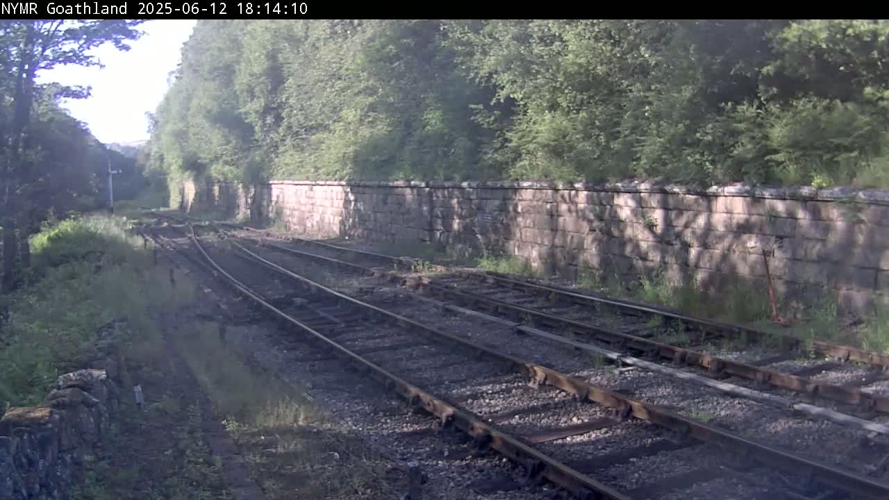 A view of multiple train tracks running alongside a stone wall, bordered by lush green vegetation under a partly sunny sky.