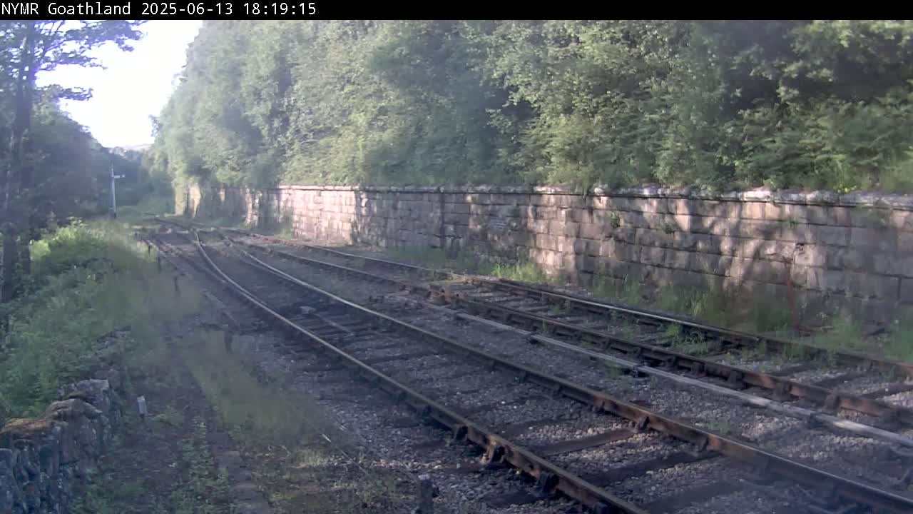 Train tracks run through a wooded area, alongside a low stone wall, under partly sunny conditions.