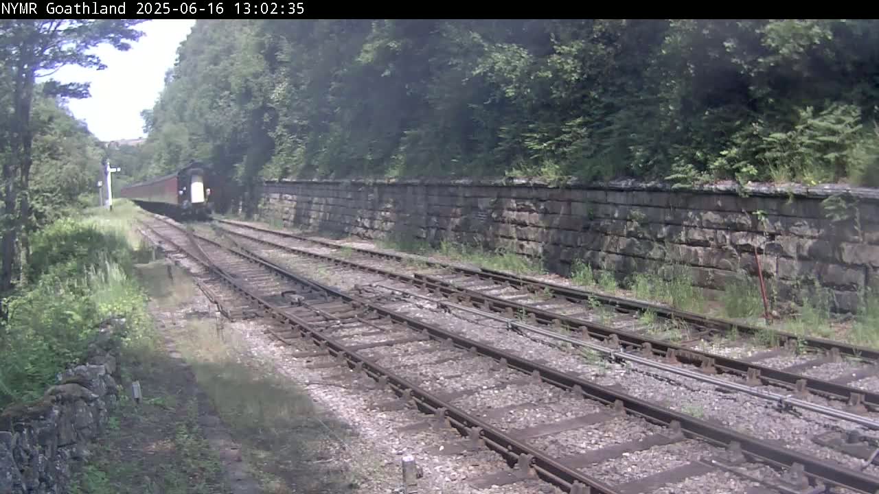 A train is traveling away from the camera along a railway track that is bordered by a stone wall and lush green vegetation on a partly cloudy day.