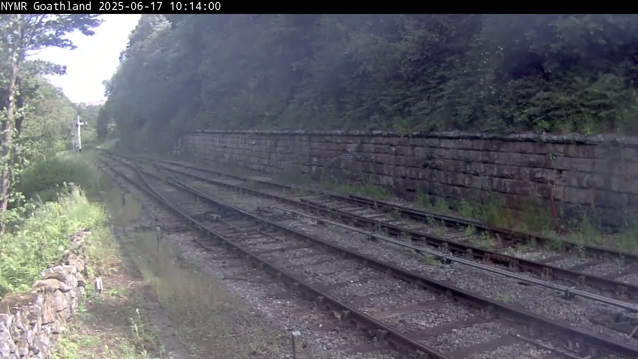 A view of a railway track running alongside a stone wall, bordered by lush greenery under hazy, overcast conditions.