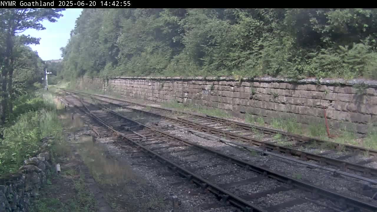 A view of a railway track bordered by a stone wall and lush green vegetation on a sunny day.