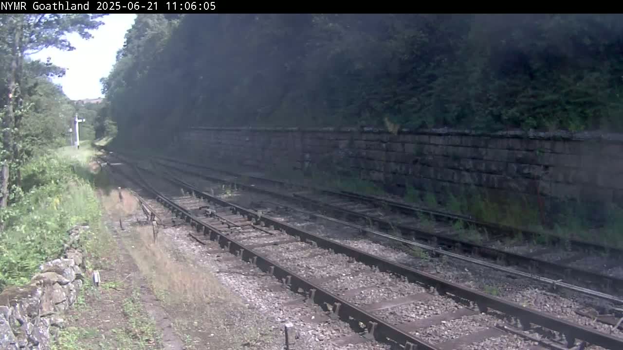 Multiple train tracks run alongside a stone wall bordered by lush green vegetation on a partly sunny day.