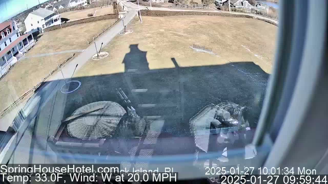 A view from a window shows a brown, grassy area with a building and its shadow, some landscaping, and a large round object in the foreground, all under a sunny sky with a 20 mph west wind.