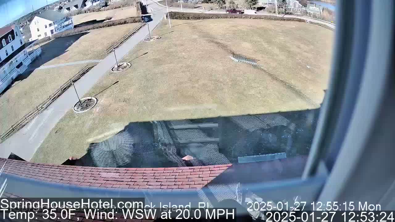 A view from a window shows a brown grassy area with a paved path and buildings in the background, under a sunny sky with a 20 mph wind.