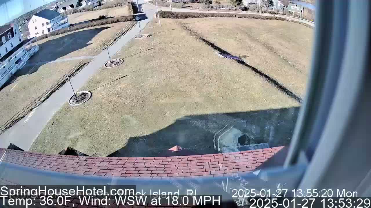 A high-angle view shows a brown, grassy area with a paved pathway, some small circular gardens, and buildings in the background on a sunny day with a WSW wind at 18 mph.