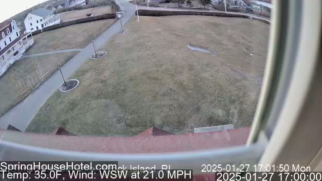 A mostly brown, grassy field is seen from a window, with a paved path, buildings, and a WSW wind at 21.0 mph.