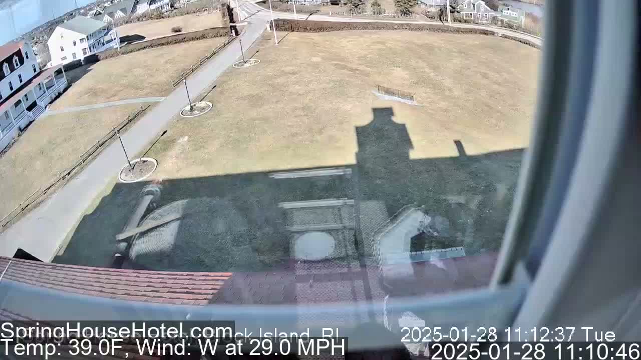 A view from a window shows a mostly brown, grassy field with a paved path, buildings in the background, and a strong west wind.