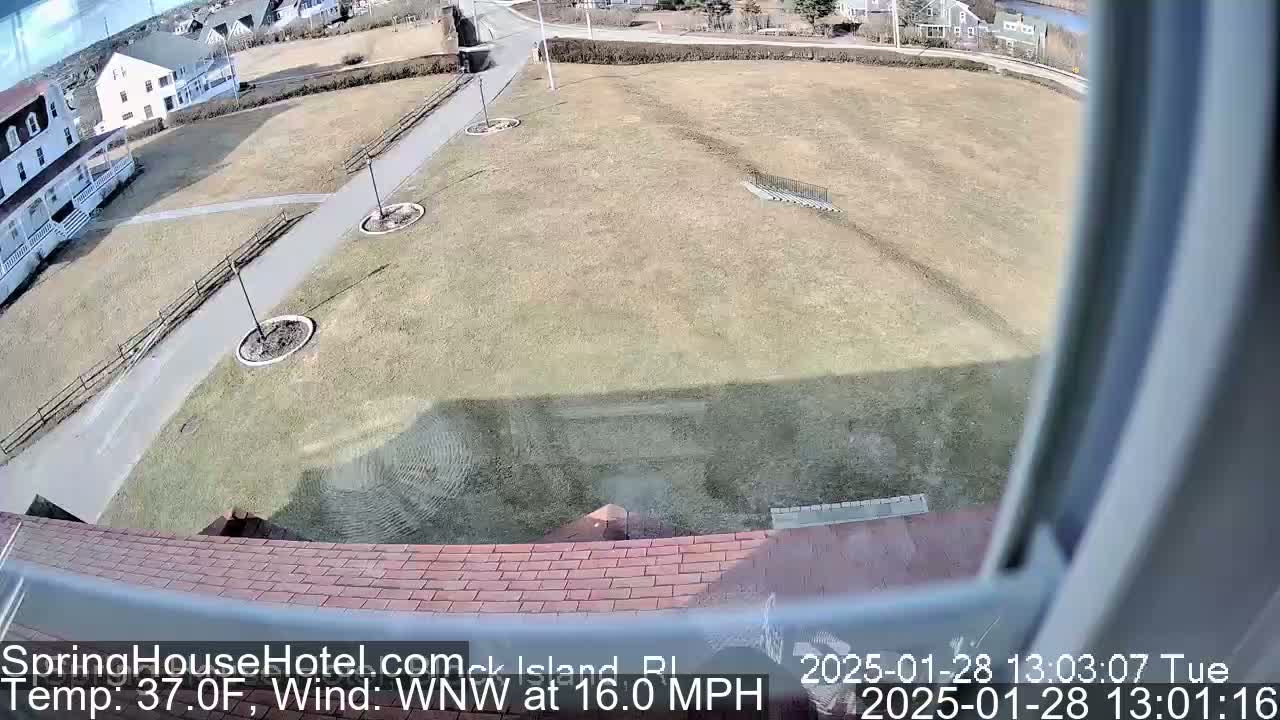 A view from a window shows a brown-roofed building overlooking a grassy area with a paved path, some landscaping, and other buildings in the distance, under sunny conditions with a WNW wind at 16 mph.