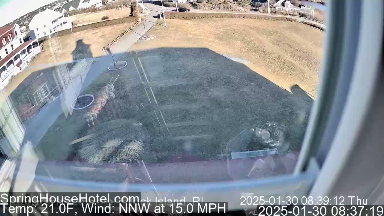 A view from a window shows a partly sunny day with a 15 mph northwest wind, overlooking a mostly barren brown lawn, several buildings, and some landscaping.