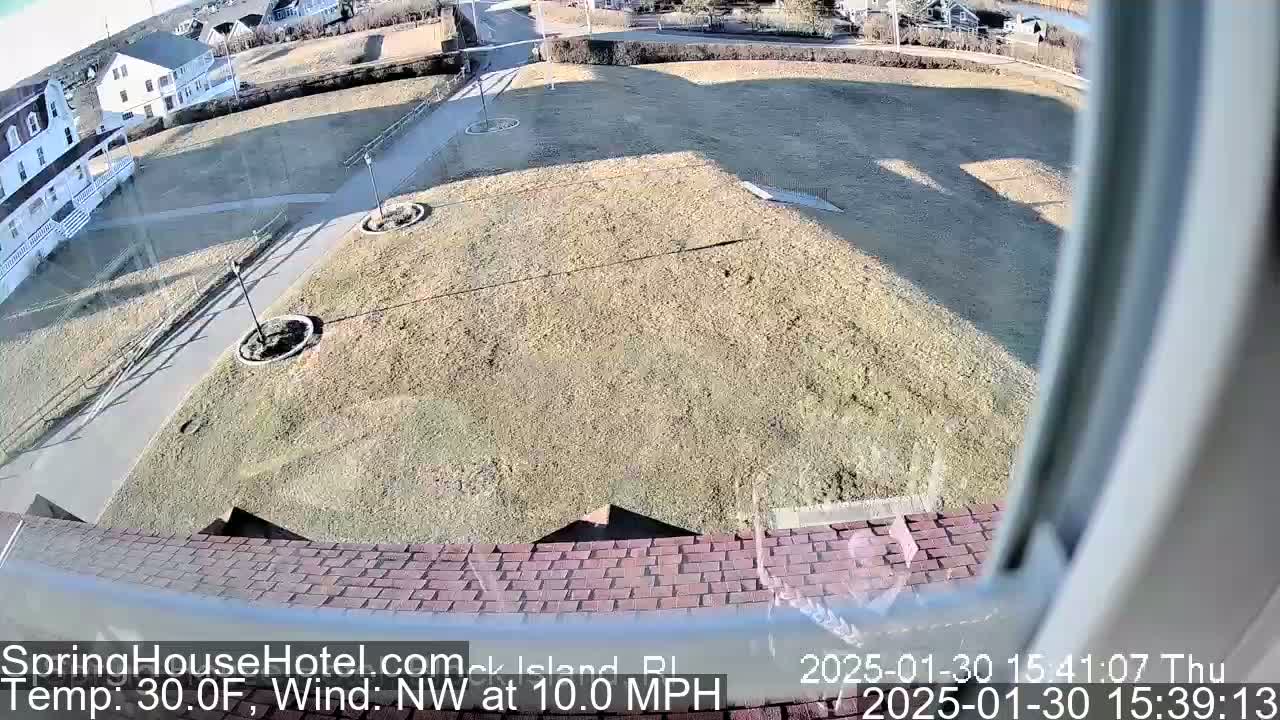 A view from a window shows a brown, grassy area with a paved walkway, some landscaping, and buildings in the background, under sunny, cold conditions with a northwest wind of 10 mph.
