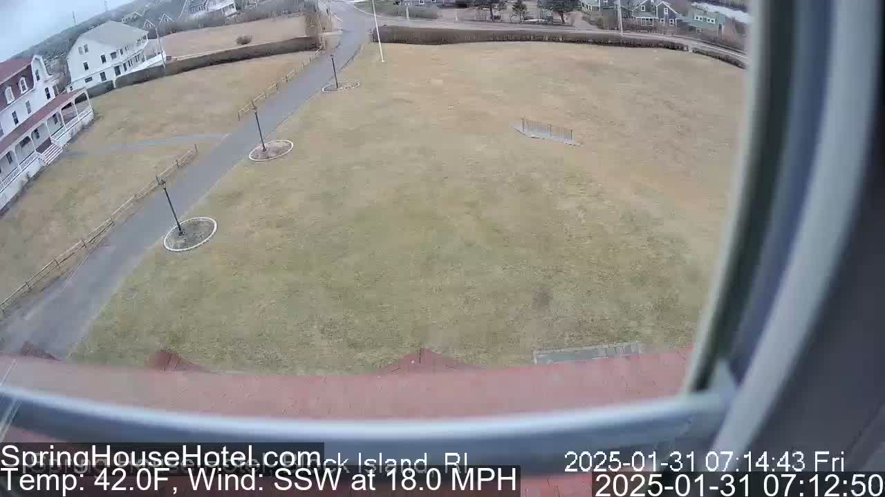 A high-angle view from a window shows a brown grassy area with a paved path, bordered by buildings and under a cloudy sky with a 18 mph southwest wind and a temperature of 42°F.