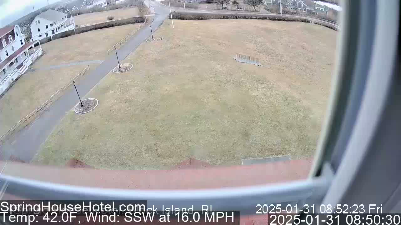 A view from a window shows a large, flat grassy area with paved walkways, lampposts, and buildings in the background under overcast skies.