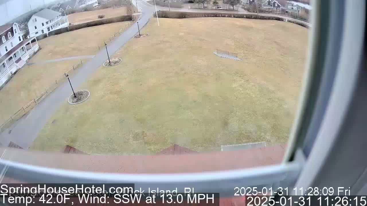 A view from a window shows a large, flat grassy area with paved walkways and buildings in the background, under a cloudy sky with a south-southwesterly wind blowing at 13 mph and a temperature of 42°F.