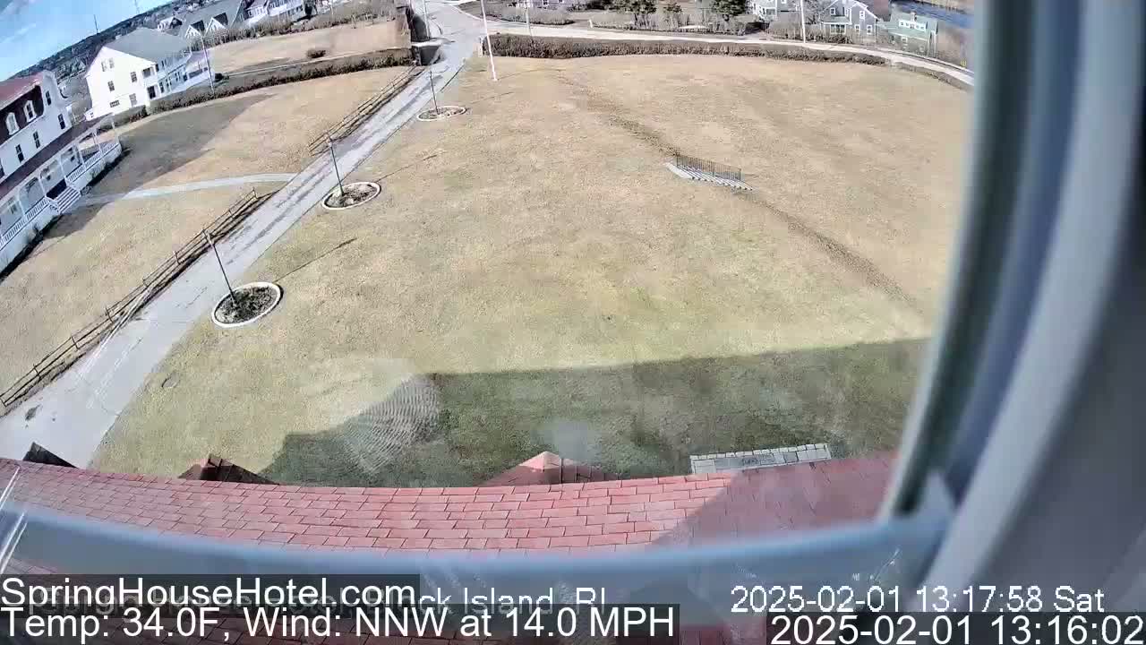 A high-angle view shows a brown, grassy field with a few paved walkways and buildings in the background, under a clear sky with a northwest wind at 14 mph and a temperature of 34°F.