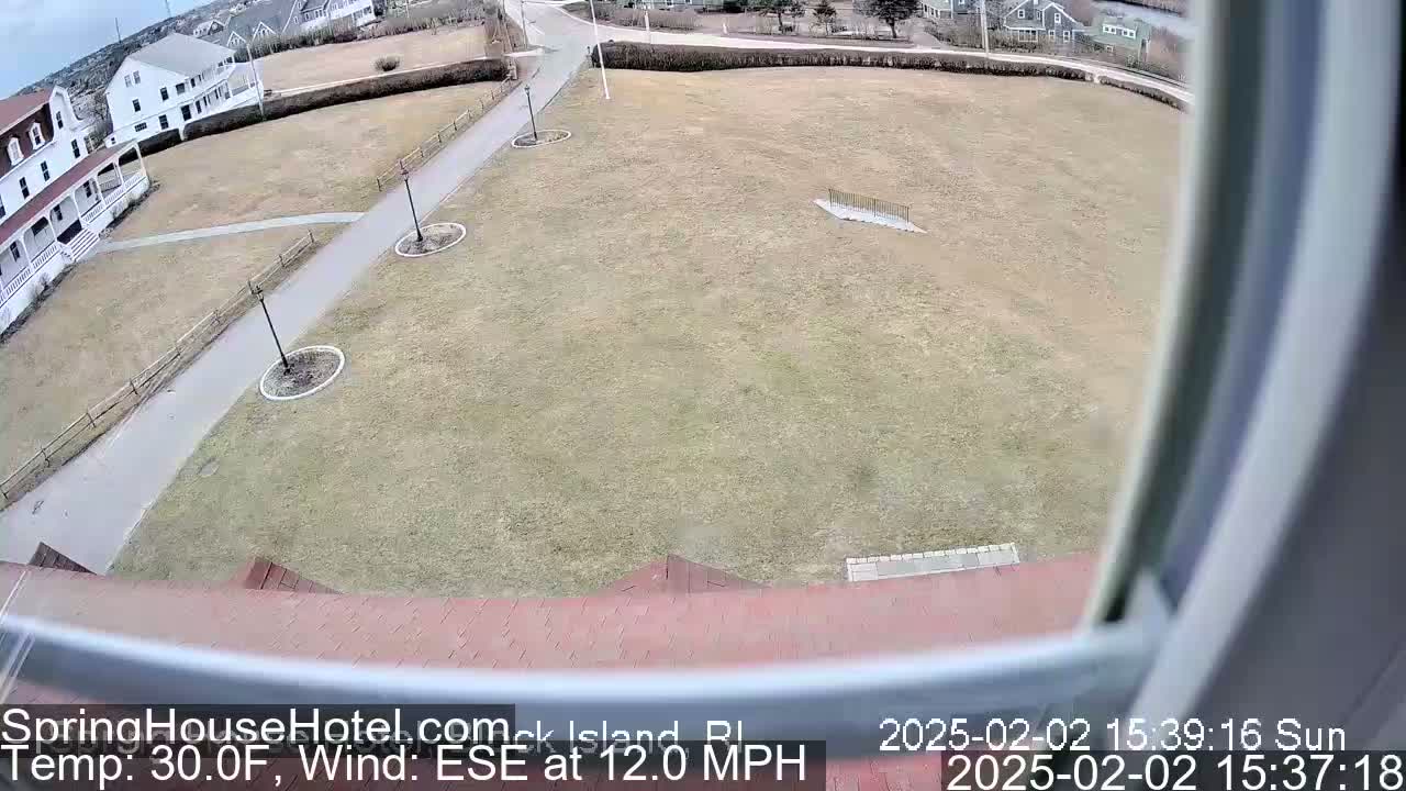 A sunny, high-angle view shows a large, mostly-brown grassy area with a paved path and some buildings in the background, under a partly cloudy sky with a 12 mph wind from the east-southeast.