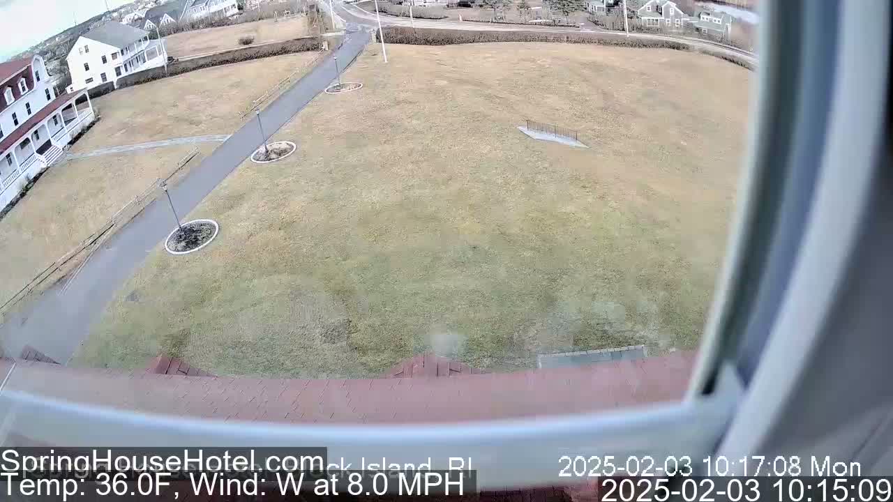 A view from a window shows a large, brown, grassy area with a paved path and several small, circular planting beds, all under overcast skies.