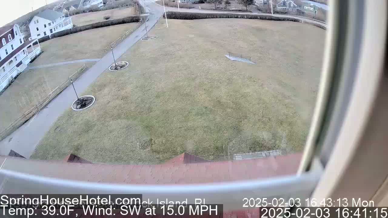 A view from a window shows a grassy area with paved pathways, landscaping, and buildings in the background, under a cloudy sky with a southwest wind at 15 mph.