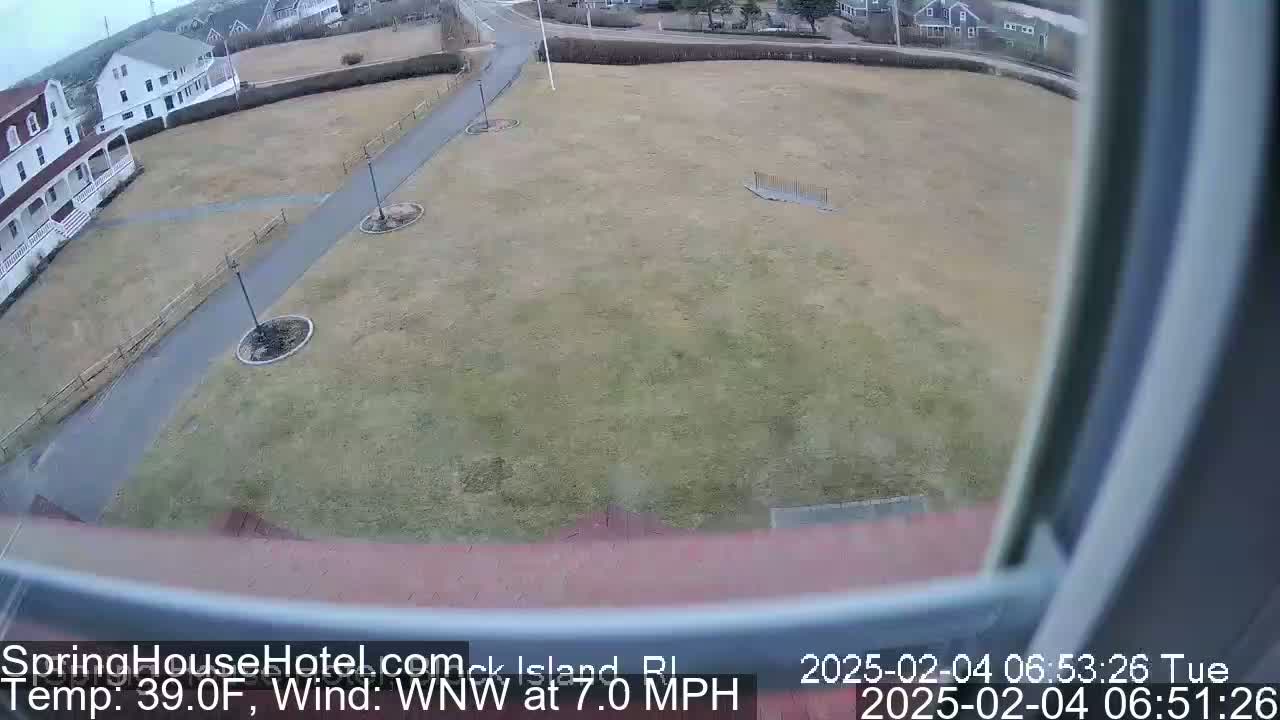 A view from a window shows a grassy field with paved pathways, some buildings, and overcast weather conditions.