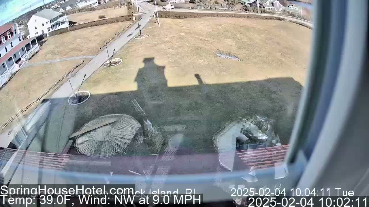 A view from a window shows a brown grassy area with a pathway, buildings in the distance, and a northwest wind at 9 mph and a temperature of 39°F.