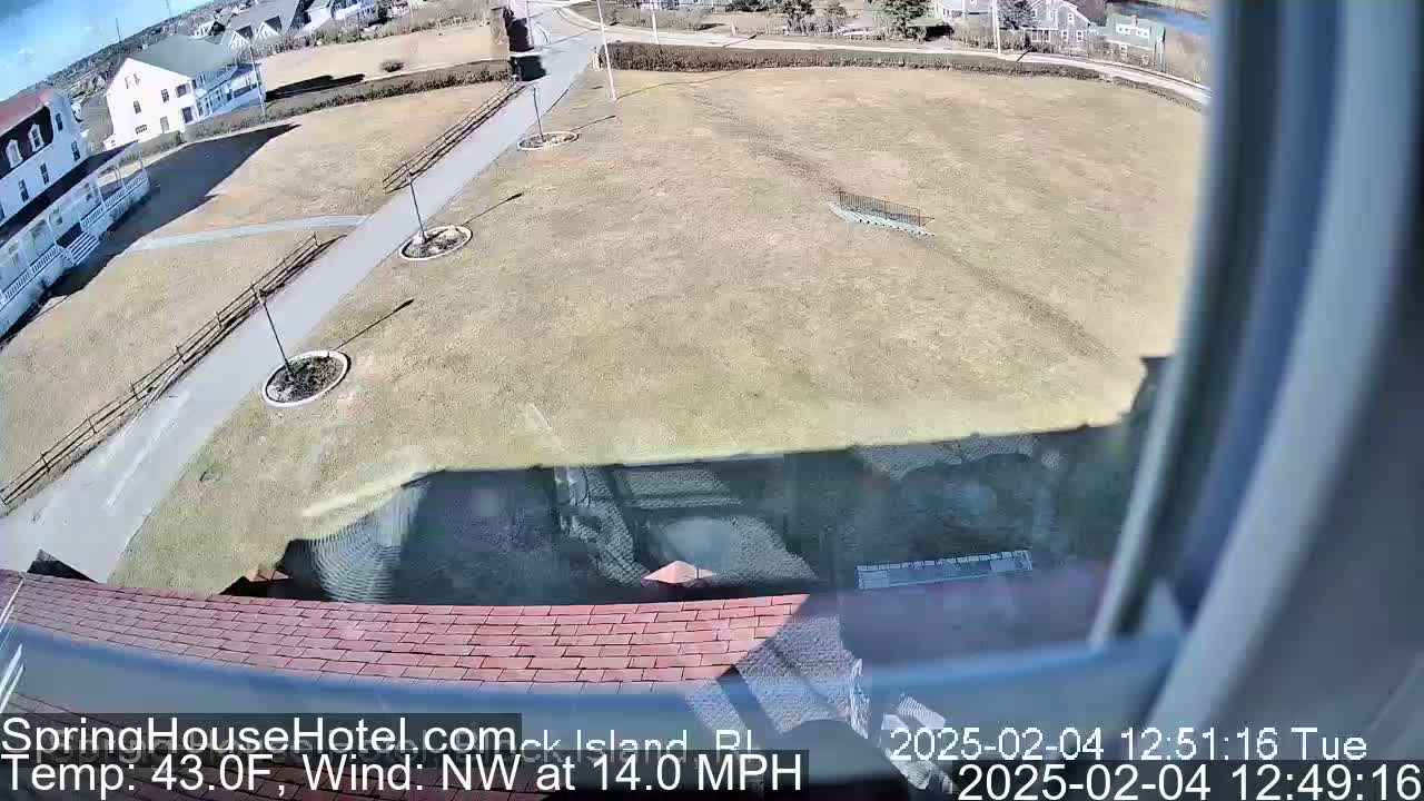 A view from a window shows a brown grassy area with a paved path, some landscaping, and buildings in the background under a sunny sky with a northwest wind.
