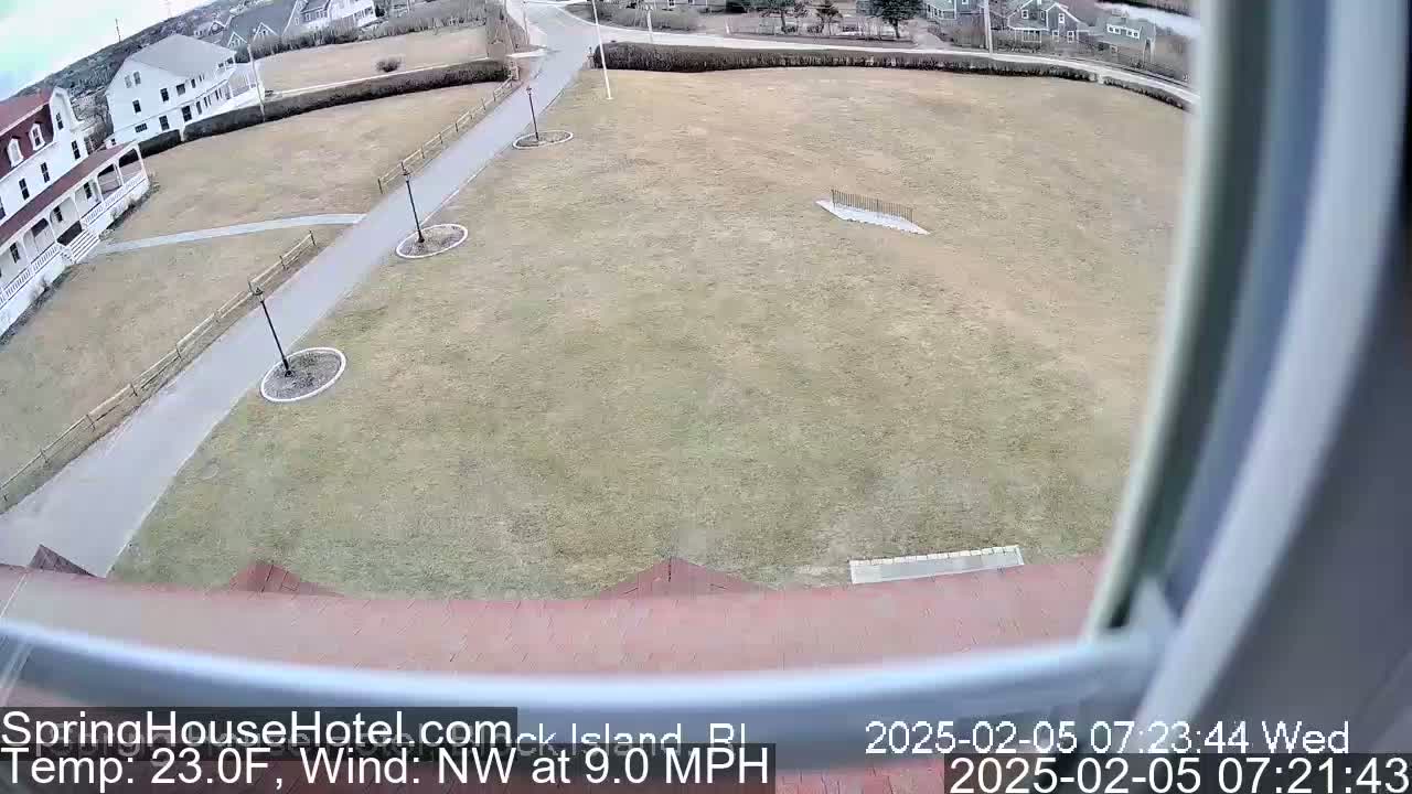 A mostly clear view of a brown grassy field with a paved walkway and some buildings in the background, under a partly cloudy sky with a northwest wind at 9 mph.
