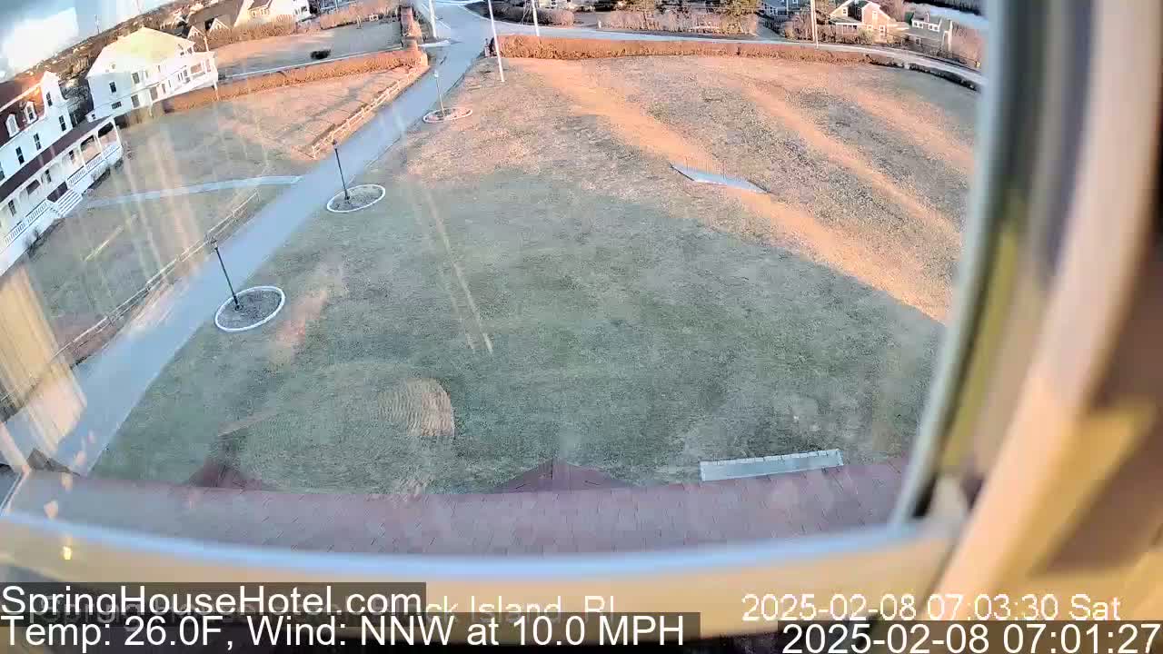 A view from a window shows a grassy area with walkways and a building, under a partly sunny sky with a north-northwest wind at 10 mph and a temperature of 26 degrees Fahrenheit.