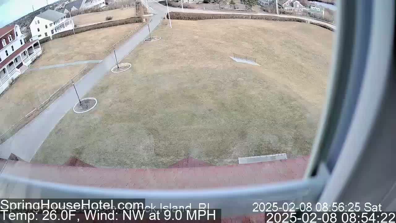 A view from a window shows a brown, grassy field with a paved pathway, a building, and a northwest wind at 9.0 mph.