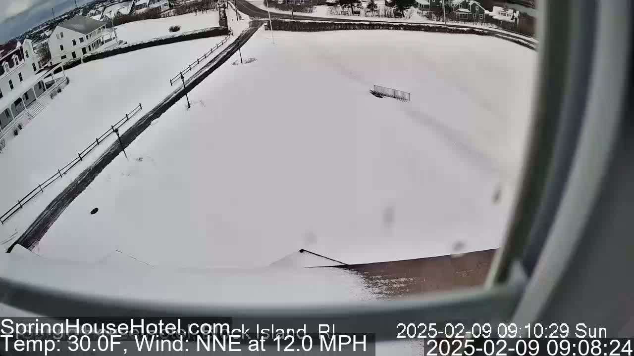 A snow-covered landscape is viewed from an elevated position, showing a road, buildings, and a fence under a sunny sky with a north-northeast wind at 12 miles per hour and a temperature of 30 degrees Fahrenheit.