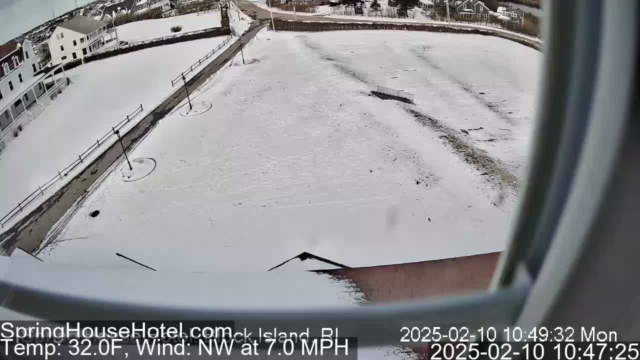 A snow-covered field with a road and buildings in the background, viewed from above on a 32°F day with a northwest wind at 7.0 mph.