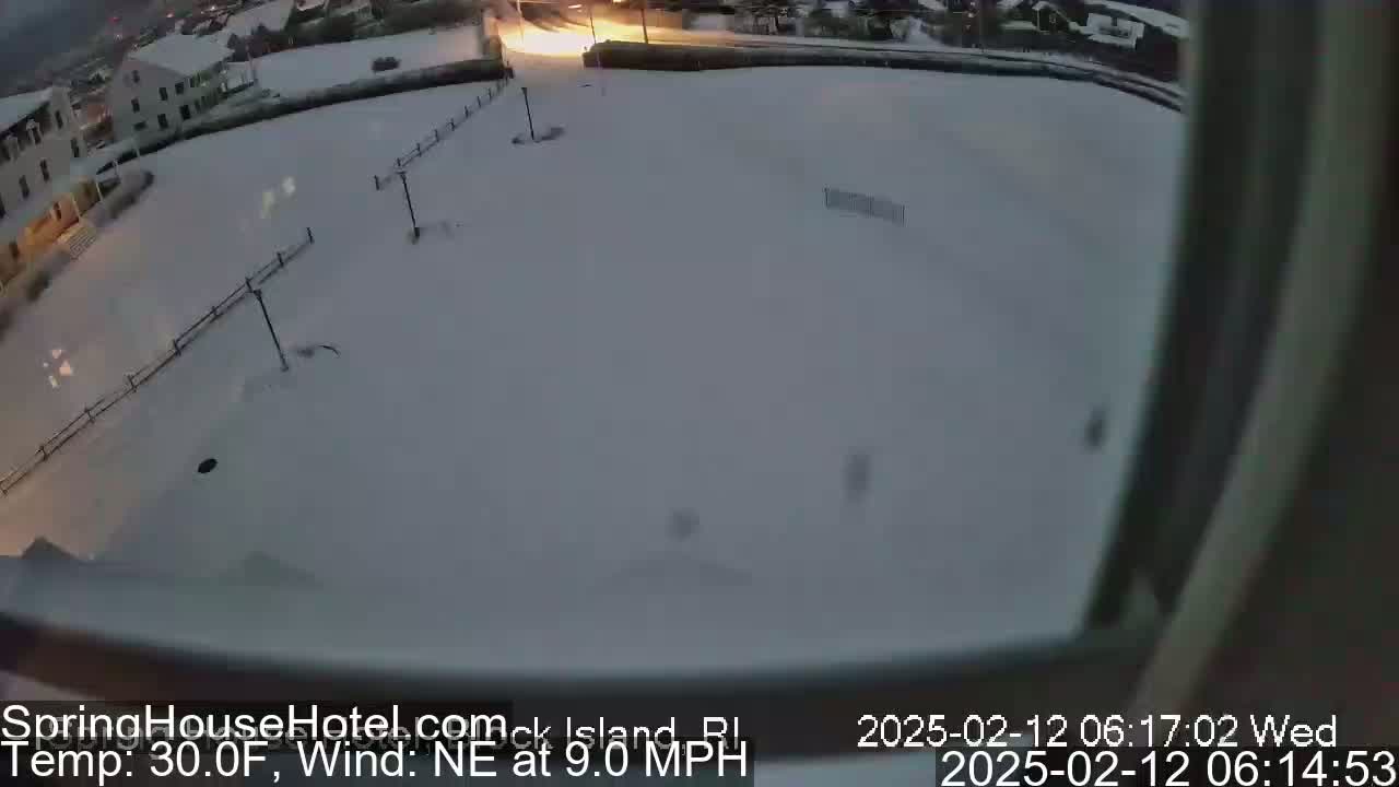 A snow-covered ground viewed from a window, with a building and some streetlights visible in the distance, under a dark sky.