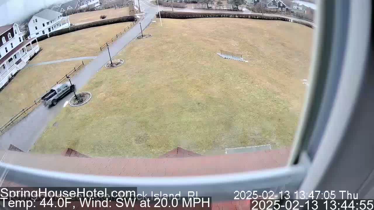 A pickup truck drives on a wet road past a grassy area with buildings in the background, under a cloudy sky with a southwest wind blowing at 20 mph.