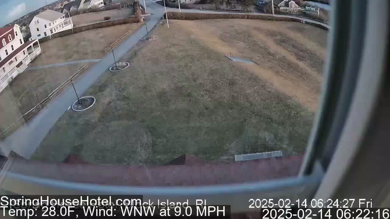 A view from a window shows a mostly brown, grassy area with a paved walkway, a few lampposts, and a building in the background, under partly sunny skies.