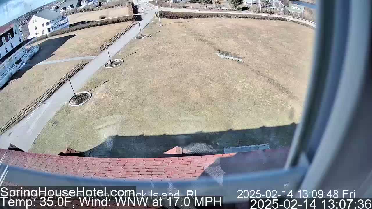A view from a window shows a brown grassy field with a paved path, some landscaping, and a building in the background under sunny, windy conditions.